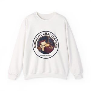 Op de afbeelding: Witte sweatshirt met een cirkelvormige afbeelding van een slaapkamer. De afbeelding toont een bed, een open boek, een kat en de tekst "MIDNIGHT CHAPTER CLUB" en "lifetime member."