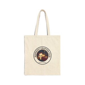 May include: A cream-coloured tote bag with a circular graphic depicting a bedroom scene. The graphic includes the text "MIDNIGHT CHAPTER CLUB" and "lifetime member." The bag has two long handles and is made of a sturdy fabric.
