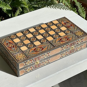 Puede incluir: Un tablero de juego de backgammon de madera con intrincados diseños de incrustaciones en tonos marrones, rojos y dorados. El tablero está cerrado y presenta un patrón geométrico tradicional.
