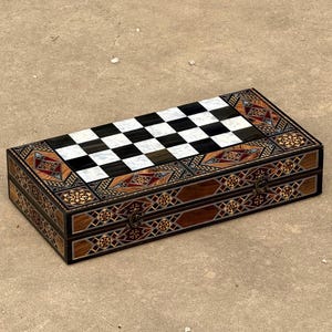 May include: A rectangular wooden chess set box with a checkered playing surface. The box features intricate inlaid patterns in various colors, including brown, black, white, and red. The box has a clasp closure and is designed for chess.