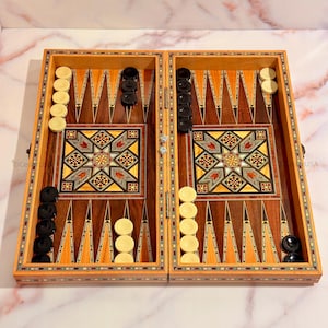 May include: A wooden backgammon board with inlaid designs. The board is open and shows the playing surface with black and white checkers. The board is made of dark wood with a light wood inlay.