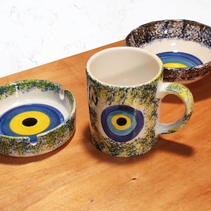 May include: A set of three ceramic dishes with a blue and yellow evil eye design. The dishes are a mug, a small bowl, and an ashtray.