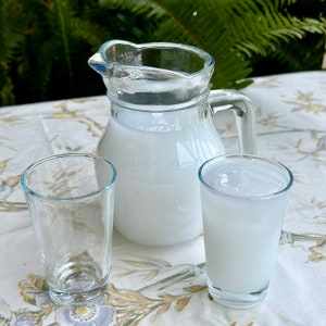 May include: A glass jug and two glasses filled with a white, cloudy liquid, possibly a traditional Greek drink like ouzo or raki, on a floral tablecloth.