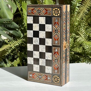 Puede incluir: Un tablero de ajedrez de madera con una superficie de juego de cuadros negros y blancos. El tablero está incrustado con intrincados patrones geométricos en tonos rojos, marrones y dorados. El tablero está cerrado y tiene un cierre de latón.