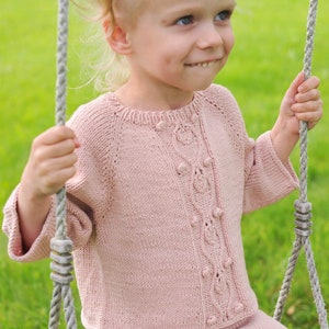 May include: A young girl wearing a pink knitted cardigan with a button detail sits on a swing. The cardigan has a cable knit pattern and is made with a soft yarn.