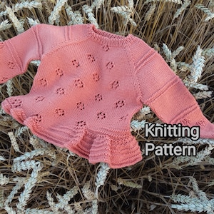 May include: A pink knitted baby sweater with a lacy pattern and a ruffled hem. The sweater is laid out on a bed of dried wheat stalks. The text "Knitting Pattern" is visible in the lower right corner of the image.