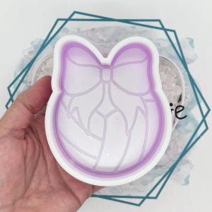 May include: A white and purple volleyball-shaped cookie cutter. The cutter features a bow design and a volleyball pattern. The cutter is held in a hand.
