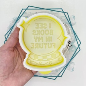 May include: A yellow crystal ball-shaped cookie cutter with the text "I SEE BOOKS IN MY FUTURE". The cutter has a white border and star accents. The design is on a light blue and white background.