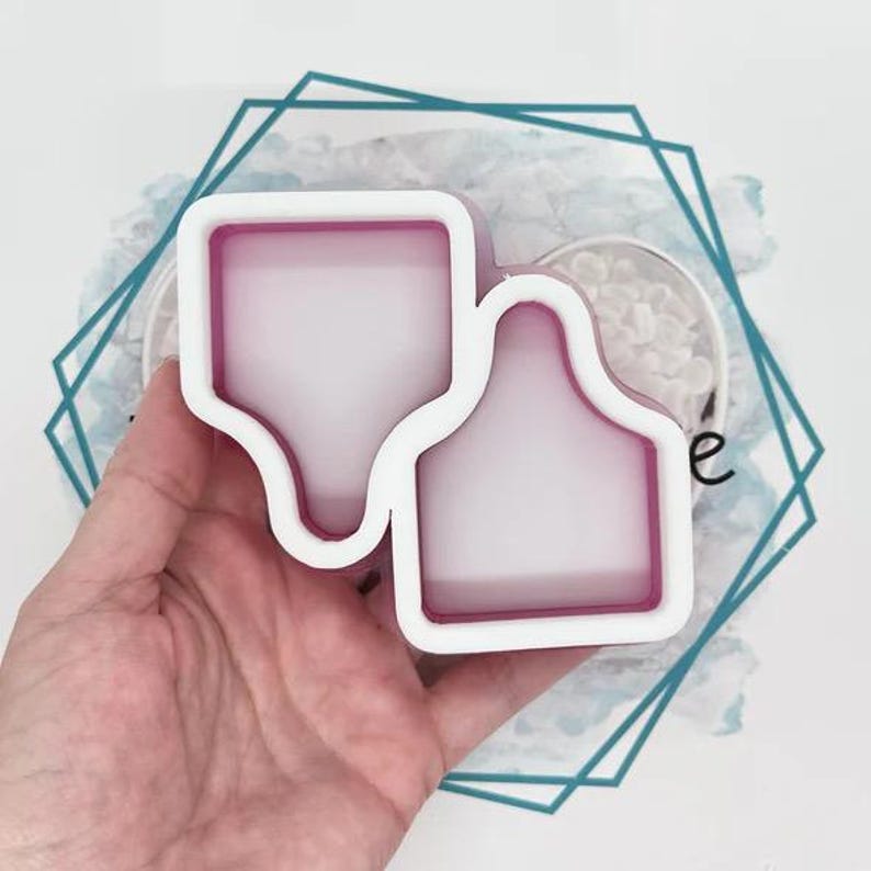 May include: Two white and pink silicone moulds for crafting, featuring a distinct tag-like shape with a flat bottom and a rounded top section. The moulds have a smooth, translucent interior surface and a thick, opaque white outer border frame.