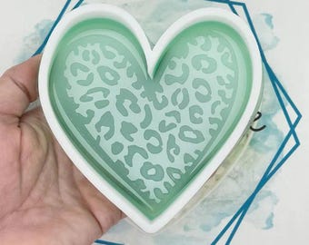 May include: A heart-shaped mold with a white border and a light green interior. The interior features a leopard print design. The mold is held by a hand, and the background includes a blue geometric shape.
