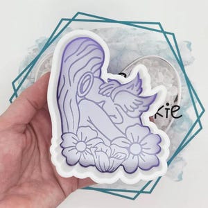 May include: A white and purple silicone mould featuring a detailed illustration of a figure with wings, flowers, and long hair. The design is in a gradient of purple hues. The mould is held in a hand.