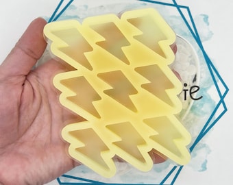 May include: A yellow silicone mold with nine lightning bolt shapes. The mold is held in a hand and is on a white surface with a blue geometric design in the background. The lightning bolt shapes are evenly spaced.