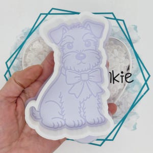May include: A light purple cookie cutter shaped like a sitting dog with a bow around its neck. The cutter has a thick white plastic border and a detailed embossed outline of the dog's fur, facial features, and bow on the interior surface.