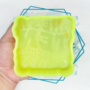 May include: A neon green silicone mould with the text "SHE GETS THE HITS 28" in a retro font. The mould has a square shape with a wavy border. The mould is held in a hand.