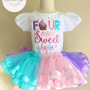 May include: A white short-sleeved shirt with the text "FOUR ever sweet hayden" and a cupcake graphic. The shirt is paired with a multi-layered tulle skirt in shades of pink, blue, and purple.