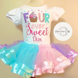 May include: A white t-shirt with the text "Four Ever Sweet" and a cupcake graphic. The shirt is paired with a multi-colored tulle skirt with pink, blue, and purple layers.