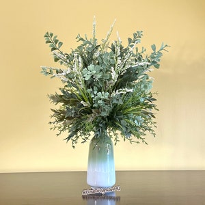 May include: A decorative floral arrangement in a green and white ombre vase. The arrangement features a variety of green foliage and white accents, creating a natural and fresh aesthetic. The vase is cylindrical with a wide base.