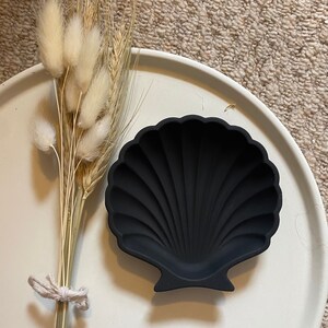 May include: A black seashell-shaped dish on a white surface. The dish is made of a smooth, matt material and has a slightly raised lip.