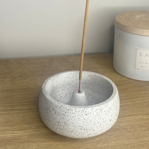 May include: A white speckled ceramic incense holder with a cone-shaped incense burner. An incense stick is burning in the holder.