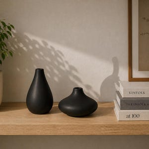 Może przedstawiać: Dwa matowe czarne wazony ceramiczne o różnych kształtach stoją na jasnej drewnianej półce. Jeden ma kształt łzy, drugi jest okrągły. Widoczna jest również roślina doniczkowa i oprawione dzieło sztuki.