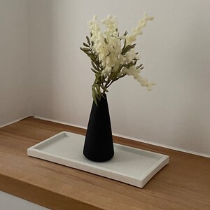 May include: A black, cone-shaped vase holds a bouquet of white flowers with green stems. The vase sits on a rectangular, light gray tray. The tray rests on a wooden surface, against a white wall.