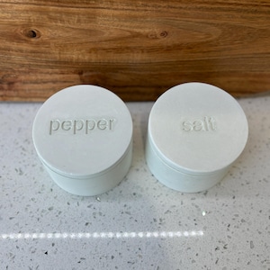 Könnte beinhalten: Zwei weiße Salz- und Pfefferstreuer aus Keramik mit den Worten "Salz" und "Pfeffer" auf der Oberseite.