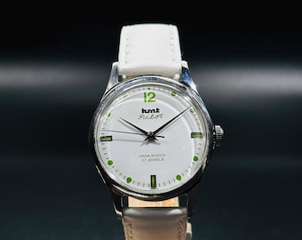 May include: A vintage wristwatch with a white face, silver case, and white leather band. The watch face features green accents, black hands, and the text "hmt Pilot." The watch is displayed on a wooden stand.