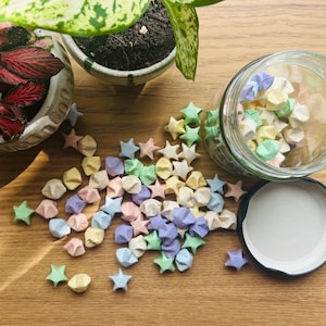 May include: A glass jar filled with pastel coloured origami stars. The jar is sitting on a wooden surface with green plants in the background.