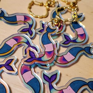 May include: A collection of mermaid-shaped keychains. Each keychain features a mermaid with a blue scaled tail, a pink midsection, and a purple tail fin. The keychains have gold-colored metal rings.
