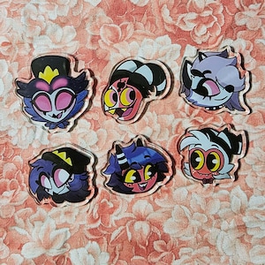 May include: Six acrylic pins featuring stylised cartoon character heads from Helluva Boss. The collection includes Stolas, Blitzø, Loona, Octavia, Millie, and Moxxie, each rendered with vibrant colours, expressive facial features, and distinct headwear.