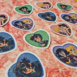May include: A collection of heart-shaped stickers featuring cartoon illustrations of two characters with dark hair and blonde hair. The stickers display various expressions and poses, with some showing the characters together and others showing them alone.