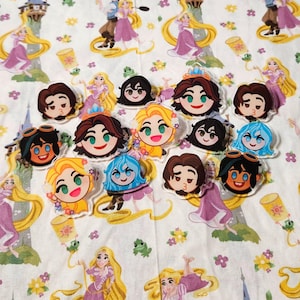 Pode incluir: Uma coleção de pequenos alfinetes circulares de acrílico com rostos de personagens de desenhos animados estilizados, incluindo figuras com cabelos longos loiros, escuros e azuis, dispostos sobre um fundo de tecido branco com ilustrações repetidas.