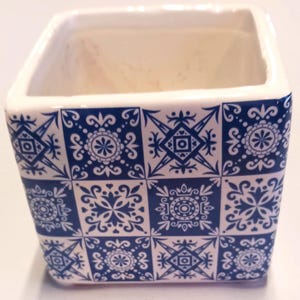 May include: A square ceramic planter with a white interior and a blue and white patterned exterior. The pattern features repeating geometric designs and floral motifs. The planter is suitable for small plants or succulents.