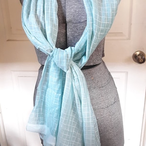 May include: A light blue scarf with a white grid pattern is draped over a gray dress form. The scarf is knotted at the front, with the ends hanging down. The fabric appears lightweight and semi-transparent.