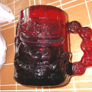 May include: A ruby red glass mug with a detailed embossed design. The mug features a handle with circular accents and a face design on the front. The mug is made of a dark red glass.