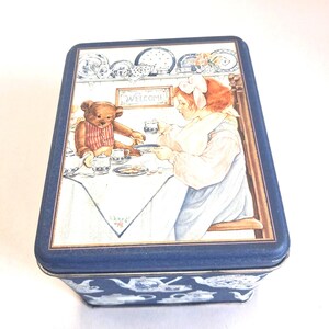 May include: A rectangular blue metal tin featuring a lid with an illustration of a person and a teddy bear having tea at a table. The scene includes a sign that reads Welcome, a tablecloth, and tea service. The sides of the tin feature a white teapot pattern.