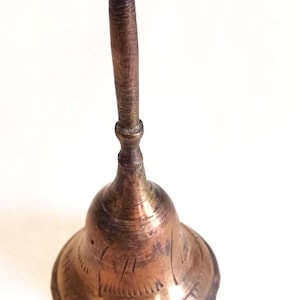 May include: A small, antique-style brass bell with a long, textured handle. The bell has etched decorative lines around the base and a slightly tarnished finish, suggesting age. The bell is approximately 5 inches tall.