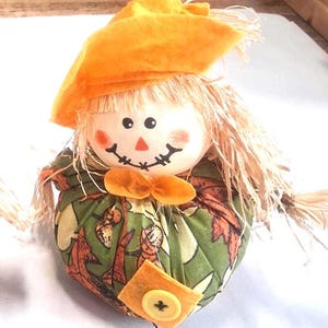May include: A scarecrow figurine featuring a round white head with a painted face, straw hair, and an orange felt hat. The body is a round fabric pouch with a green background, brown leaf patterns, an orange bow, and a yellow button.