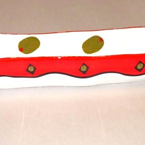May include: A white ceramic serving dish with a red border and olive decorations. The dish has an oblong shape and is decorated with green olives with red accents and black diamond shapes.
