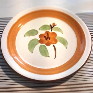 May include: A round ceramic plate with a cream-colored center and a wide orange rim. The center features a hand-painted design of a single orange flower with five petals and several light green leaves arranged around the stem on a white background.