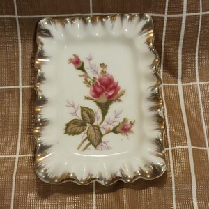 May include: A small, white porcelain trinket dish with a gold rim and a floral design featuring a pink rose and green leaves.