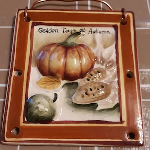 May include: A ceramic tile with a brown frame and a white background. The tile features a 3D image of a pumpkin, corn, and leaves. The text "Golden Days of Autumn" is written in brown lettering at the top of the tile.