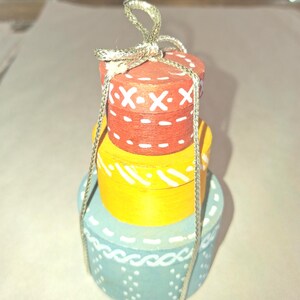 May include: A stacked, hand-painted ornament resembling a pile of gifts. The top box is red with white X's, the middle is yellow, and the bottom is blue with white accents. A silver string is attached for hanging.
