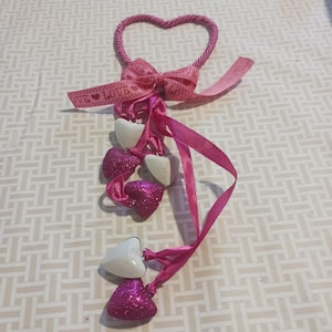 May include: A pink heart-shaped decoration with a bow and dangling hearts. The heart-shaped frame is made of twisted pink cord. A pink bow is attached to the top, with a ribbon that reads "LOVE". Dangling from the bow are white and glittery pink hearts.