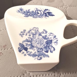 May include: White ceramic mug with a blue floral design. The mug features a curved handle and a wide, flat top. The floral pattern includes roses and other flowers.