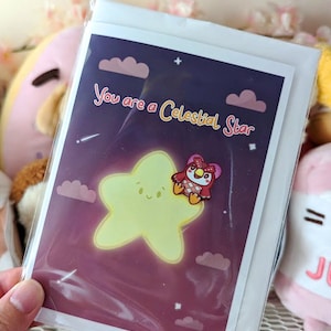May include: A greeting card with a purple and pink night sky background. A yellow star with a smiling face is in the center of the card. A small red owl with a pink bow is perched on the star. The text "You are a Celestial Star" is written in white on the card.