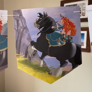 Peut inclure: Une banni&egrave;re en forme de fanion avec une illustration de dessin anim&eacute; d'un cheval noir et de cavaliers. Les cavaliers, aux cheveux boucl&eacute;s roux, portent des tenues bleues et dor&eacute;es. L'arri&egrave;re-plan repr&eacute;sente une montagne et le ciel.