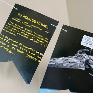 May include: A black and yellow banner with the text "THE PHANTOM MENACE" and a description of the plot of the movie. The banner is hanging from a white string. A black and grey spaceship is visible in the background.