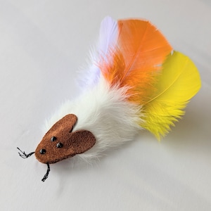 May include: A white, brown, and multi-colored feather cat toy with a brown suede mouse head. The toy has black eyes and a long tail.