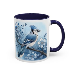 May include: White ceramic mug with a blue interior and handle. The mug features a detailed illustration of a blue jay perched on a branch with blue flowers. The design is set against a soft blue background.
